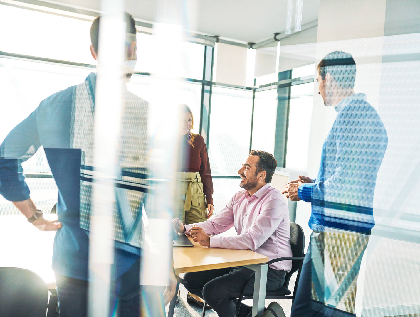 Four coworkers are talking around a desk in an office with large glass windows. Three people are standing around the desk, while one is sitting at the desk looking at a computer.