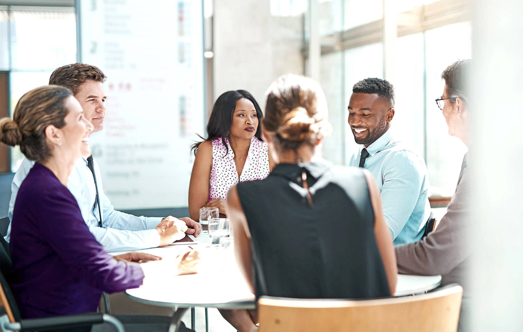 A group of six coworkers wearing business casual clothing sit at a round table. They are smiling together in an office with large glass windows.